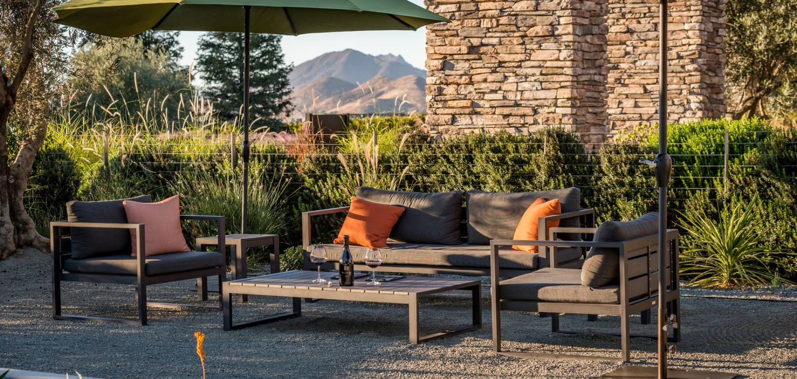 A couch and two chairs under two umbrellas at sunset with a bottle of wine on the table 