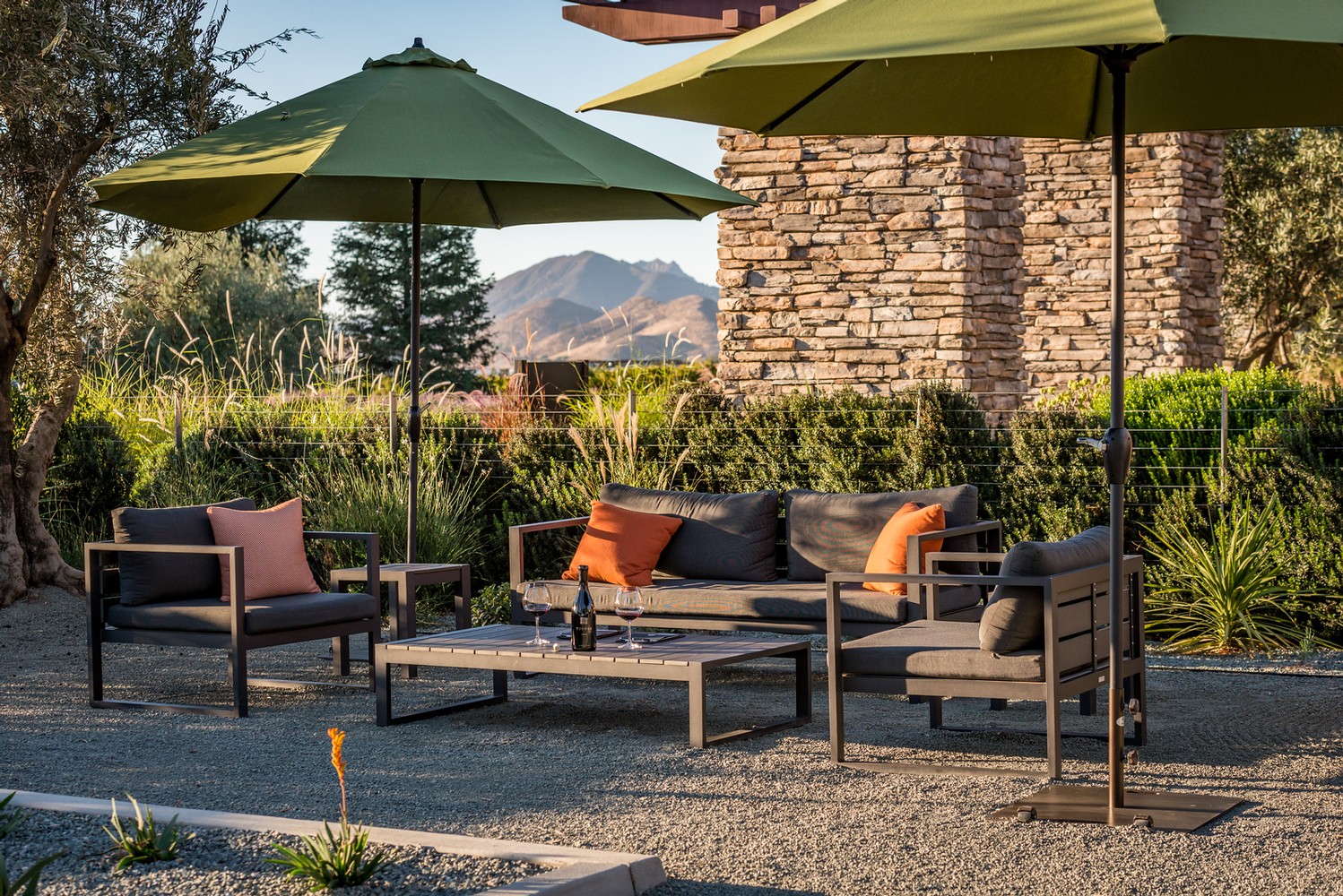 A couch and two chairs under two umbrellas at sunset with a bottle of wine on the table 