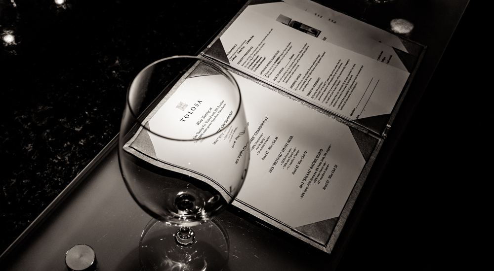 Black and white photo of a tasting room menu, with the logo reflecting through the rim of a wine glass