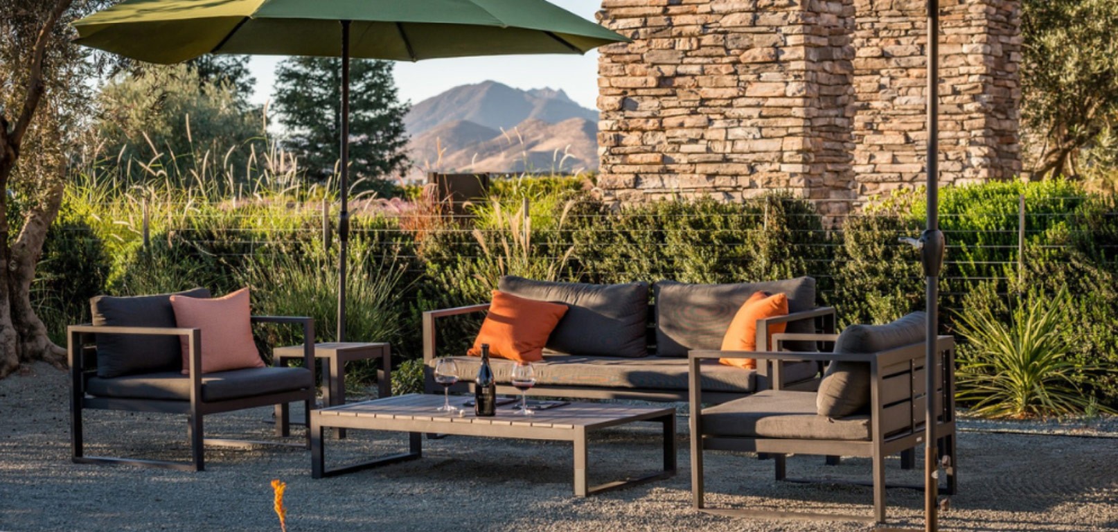 A couch and two chairs under two umbrellas at sunset with a bottle of wine on the table 