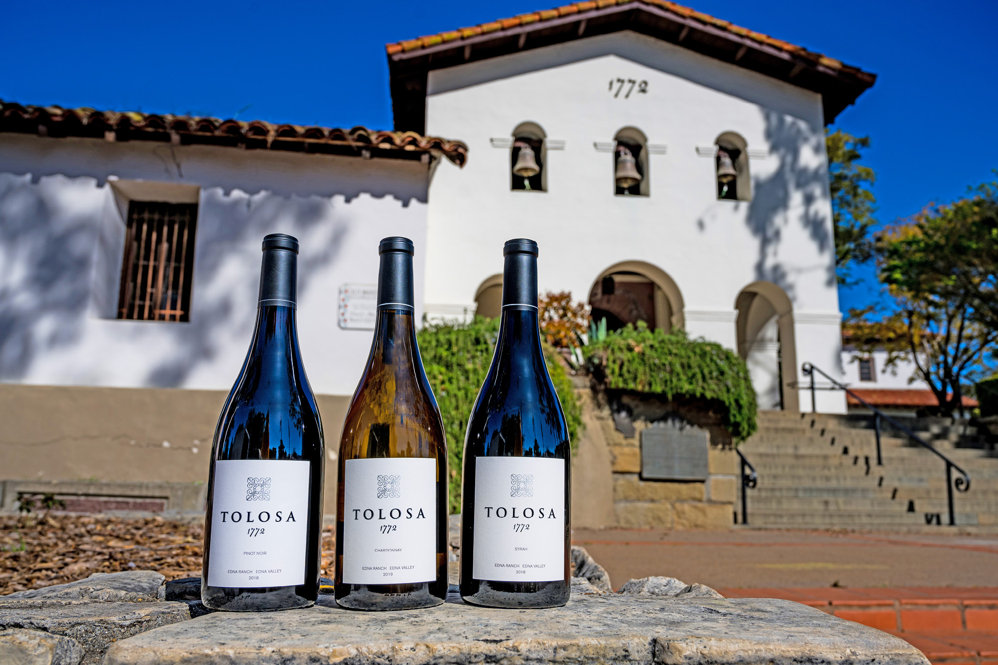 A bottle of 1772 Pinot Noir, Chardonnay, and Syrah sitting in front of the San Luis Obispo de Tolosa mission